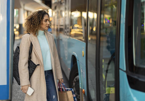 Frau mit Mobiltelefon und Einkaufstaschen am Bus - JCCMF08569