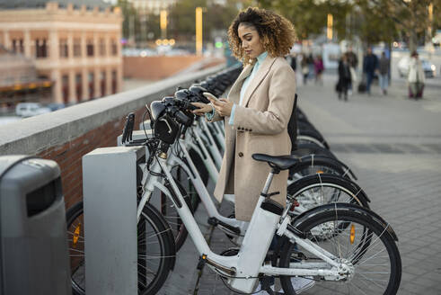 Frau mietet Elektrofahrrad über Smartphone an einer Parkstation - JCCMF08570