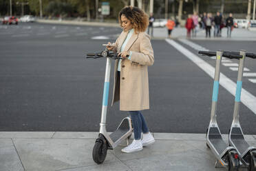 Young woman renting electric push scooter through smart phone on footpath - JCCMF08571