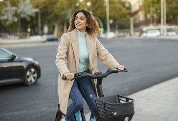 Smiling woman riding electric bicycle on footpath - JCCMF08573