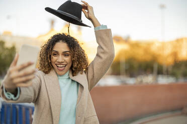 Happy woman holding hat above head taking selfie through smart phone - JCCMF08578