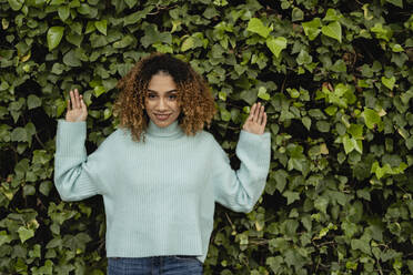 Smiling woman with curly hair leaning on plants - JCCMF08583