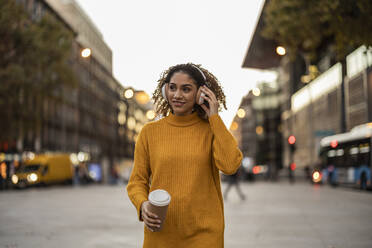 Smiling woman listening to music through wireless headphones holding disposable cup walking on footpath - JCCMF08588