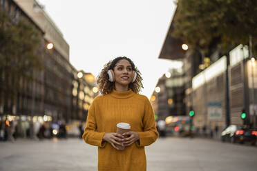 Young woman listening to music through wireless headphones holding disposable cup walking on footpath - JCCMF08589