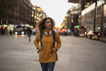 Woman with backpack and wireless headphones walking on footpath - JCCMF08594