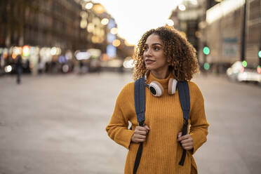 Young woman with backpack and wireless headphones walking on footpath at sunset - JCCMF08597