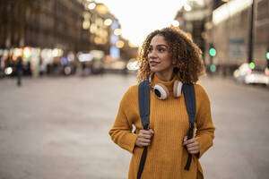 Junge Frau mit Rucksack und drahtlosen Kopfhörern, die bei Sonnenuntergang auf einem Fußweg spazieren geht - JCCMF08597