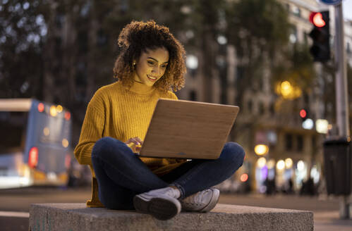 Smiling businesswoman using laptop on concrete at dusk - JCCMF08599