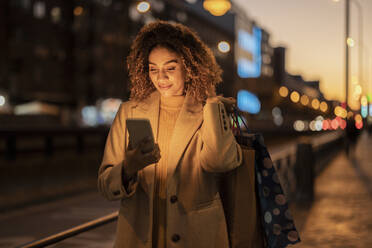 Lächelnde Frau mit Einkaufstüten und Smartphone in der Abenddämmerung - JCCMF08602