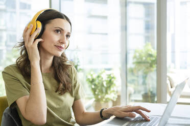 Businesswoman listening to music through wireless headphones sitting with laptop at home - SVKF00834