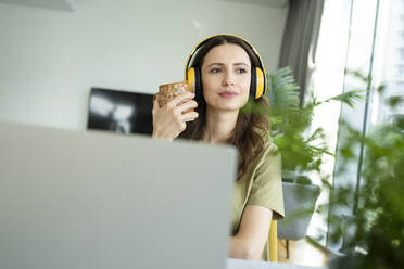Geschäftsfrau mit drahtlosen Kopfhörern, die eine Kaffeetasse in der Hand hält und durch das Fenster zu Hause hinausschaut - SVKF00836