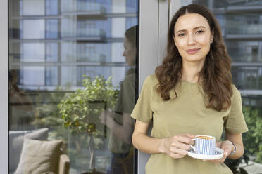 Smiling woman holding coffee cup leaning on door of balcony - SVKF00850