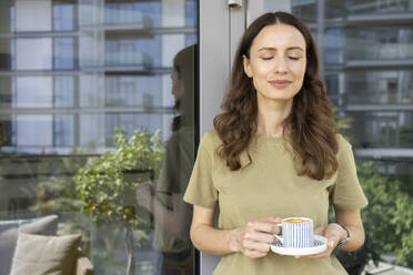 Lächelnde Frau mit geschlossenen Augen, die eine Kaffeetasse hält und sich an eine Balkontür lehnt - SVKF00851