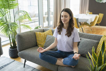 Lächelnde Frau meditiert auf dem Sofa im Wohnzimmer zu Hause - SVKF00873