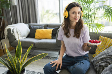 Smiling woman listening music through wireless headphones holding fresh raspberries on sofa - SVKF00876