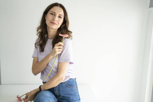 Lächelnde Frau hält Gerbera-Gänseblümchen vor einer weißen Wand - SVKF00881