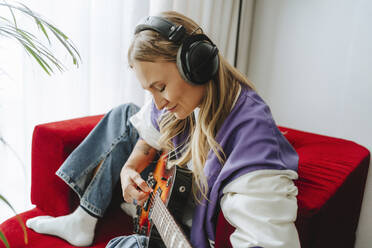 Guitarist wearing wireless headphones playing guitar on armchair - MDOF00304