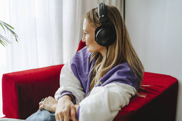 Smiling woman listening through headphones on armchair - MDOF00325