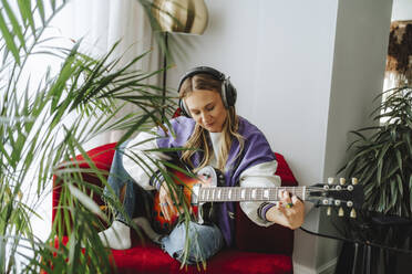 Gitarrist mit Kopfhörern spielt Gitarre auf einem Sessel im Studio - MDOF00326