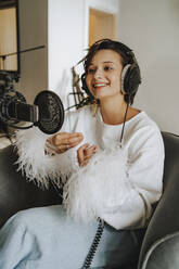 Smiling musician wearing headphones recording podcast through microphone in studio - MDOF00347