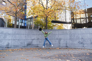 Carefree young woman holding hat jumping in front of autumn tree - ASGF03124