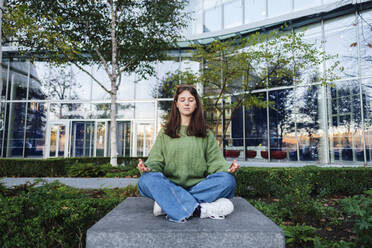 Junge Frau mit geschlossenen Augen meditiert auf einem Betonsitz vor einem Glasgebäude - ASGF03130