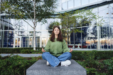 Young woman with eyes closed meditating on concrete seat in front of glass building - ASGF03130