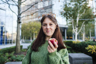 Smiling woman holding apple in front of building - ASGF03135