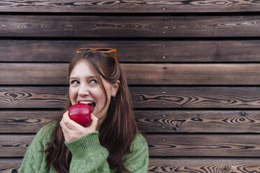 Glückliche junge Frau isst einen Apfel und lehnt sich an eine Holzwand - ASGF03136