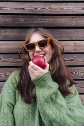 Happy woman wearing sunglasses eating apple in front of wooden wall - ASGF03137