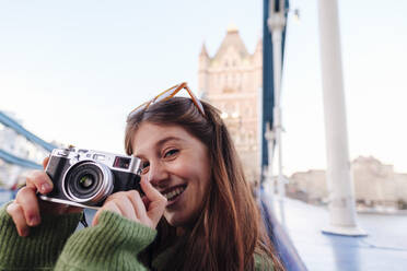 Happy young woman holding camera on Tower Bridge, London, England - ASGF03148