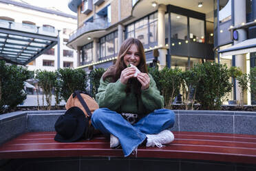 Happy young woman having sandwich on bench in front of buildings - ASGF03152