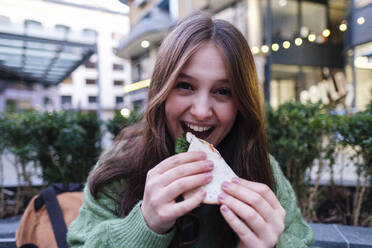 Happy young woman eating sandwich in front of building - ASGF03153