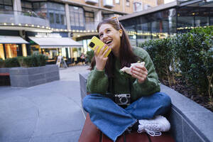 Happy woman holding sandwich talking through speaker of mobile phone on bench - ASGF03157