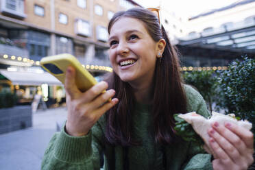 Smiling young woman holding sandwich talking through speaker of mobile phone - ASGF03159