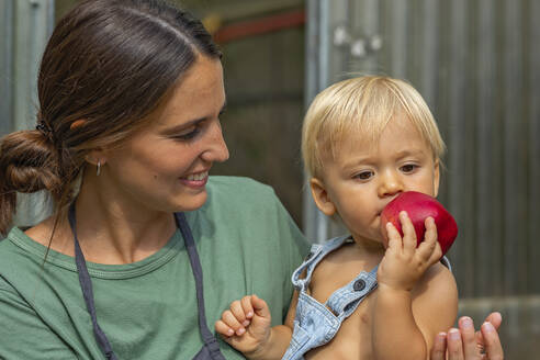 Lächelnde Mutter betrachtet ihren Sohn beim Apfelessen im Garten - MEGF00272