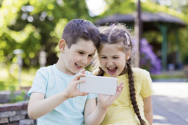 Glückliche Geschwister, die ihr Smartphone im Garten benutzen - ONAF00308
