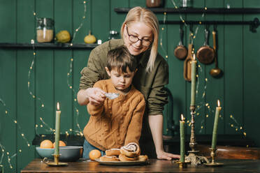 Lächelnde Mutter hilft ihrem Sohn beim Bestreuen von Zimtschnecken mit Puderzucker in der Küche - VSNF00166