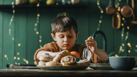 Junge hält Sieb vor Zimtschnecken und Orangen auf Tisch - VSNF00174