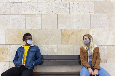 Young couple wearing protective face masks sitting on bench - JPTF01142