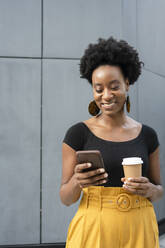 Smiling woman using mobile phone and holding disposable cup in front of wall - SVKF00893