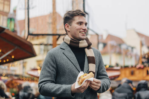 Glücklicher junger Mann mit Hot Dog auf dem Weihnachtsmarkt - VSNF00194