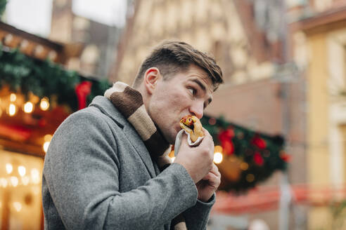 Junger Mann isst Hot Dog im Stehen auf dem Weihnachtsmarkt - VSNF00195