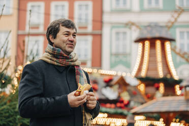 Smiling mature man standing with hot dog at Christmas market - VSNF00197