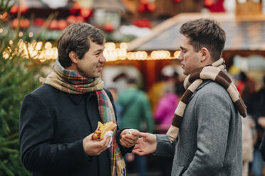 Lächelnder Vater, der einen Hotdog hält und sich mit seinem Sohn auf dem Weihnachtsmarkt unterhält - VSNF00198