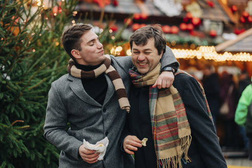 Lächelnder Mann isst Trdelnik und legt den Arm um seinen Vater, der auf dem Weihnachtsmarkt genießt - VSNF00200