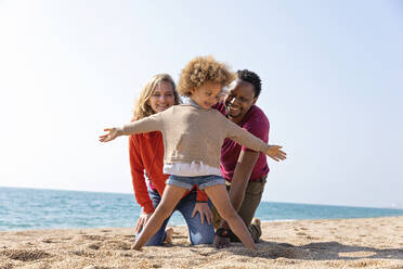 Glückliches Mädchen steht mit ausgestreckten Armen vor den Eltern am Strand - MEGF00298