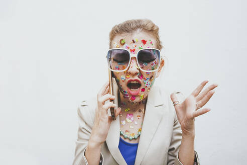 Frustrated woman with stickers on face talking on mobile phone in front of white background - SVCF00233