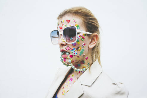 Woman with multi colored stickers on face and necklace in mouth against white background - SVCF00236