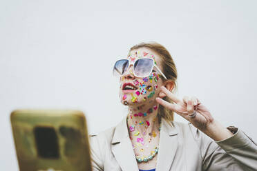 Woman with stickers on face gesturing and taking selfie through mobile phone in front of white background - SVCF00239